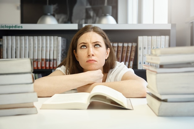 Foto gratuita studentessa caucasica arrabbiata con le sopracciglia tese che guardano su, provando a prepararsi per gli esami e leggere un manuale, avendo sguardo stanco e frustrato contro la biblioteca universitaria