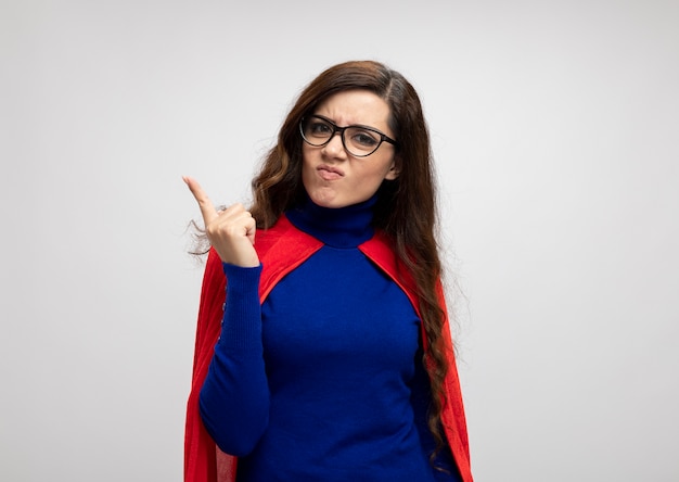 Angry caucasian superhero girl with red cape in optical glasses shows index finger isolated on white wall with copy space