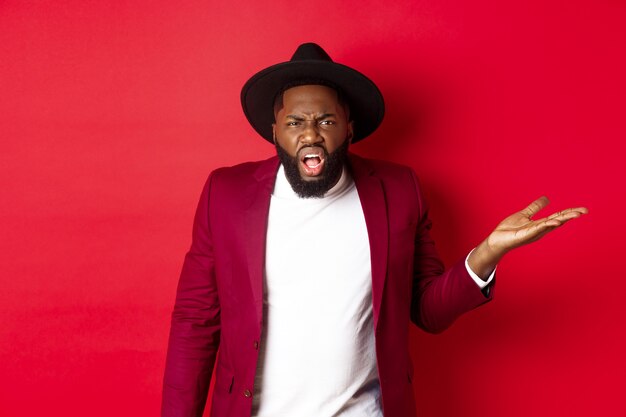 Angry and confused Black man arguing, shouting at camera and raising hand bothered, cant understand something, red background.