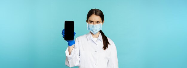 顔をしかめたスマートフォンアプリのモバイル画面を示すフェイスマスク手袋をはめた怒って混乱した女医
