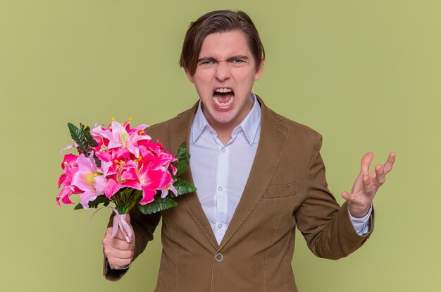 Angry crazy mad young man holding bouquet of flowers looking at front shouting going wild international women's day standing over green wall