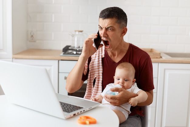 화난 검은 머리의 잘생긴 남자는 티셔츠를 어깨에 수건으로 두르고 휴대전화로 말하고 공격적인 표정으로 비명을 지르며 노트북을 들고 테이블에 앉아 아기를 손에 들고 있습니다.