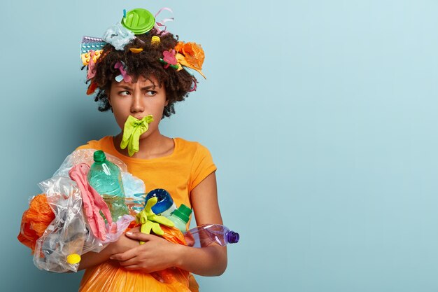 Angry dark skinned woman has rubber gloves in mouth, smirks face, being against of plastic contamination