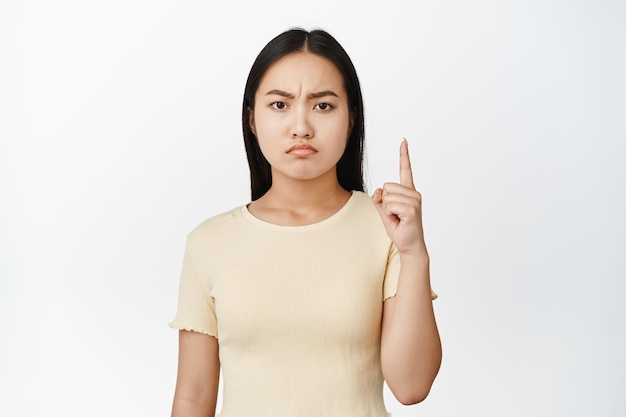 Злая и разочарованная азиатская девушка показывает пальцем вверх и обиженно смотрит в камеру, стоящую в желтой футболке на белом фоне