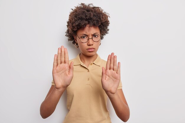 Angry displeased Afro American woman extends palms and says no shows prohibition gesture refuses something wears round spectacles casual brown t shirt isolated over grey wall