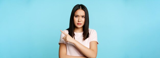 Angry displeased asian woman pointing right furrow eyebrows in disapproval standing in tshirt over b