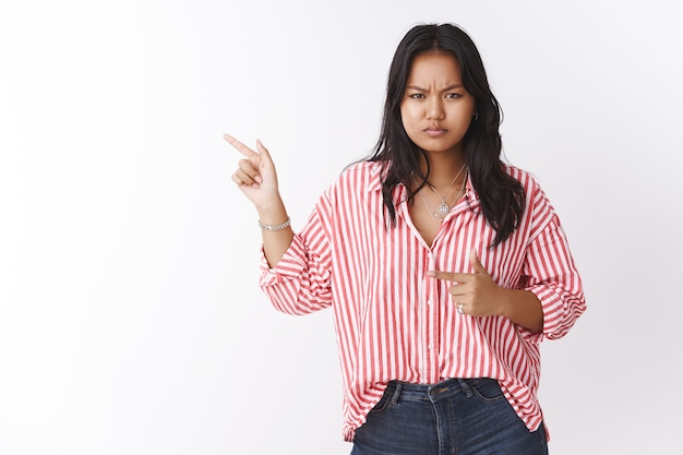 Angry displeased girlfriend asking question about girl in boyfriend house, pointing left dissatisfied and irritated frowning and squinting upset, posing frustated over white wall