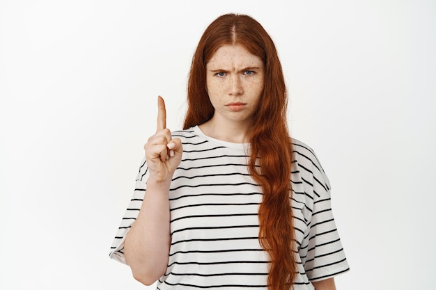 Ragazza rossa arrabbiata e dispiaciuta che punta il dito in alto giudicante, accigliata con antipatia, delusa dal banner o dal testo promozionale verso l'alto, disapprova la pubblicità, sfondo bianco