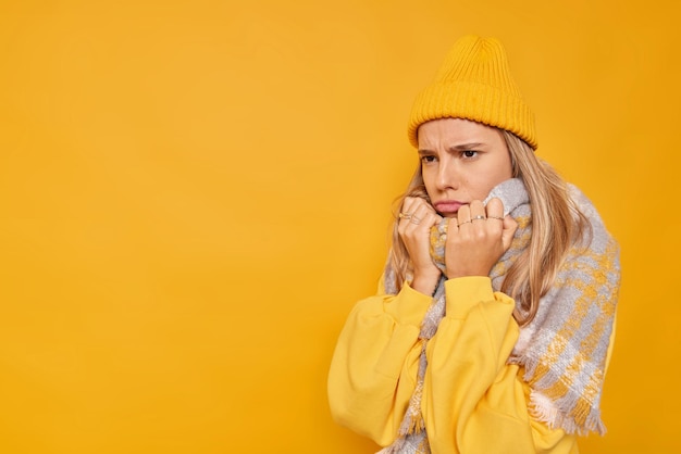 Angry displeased woman looks with sulking expression wears warm scarf around neck feels unhappy poses against yellow background blank space for your promotional content. Negative emotions concept