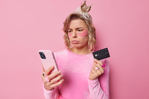 Angry dissatisfied woman with hairstyle looks attentively at smartphone screen checks her bank account upset not to have money holds credit card wears hydrogel patches under eyes poses indoor