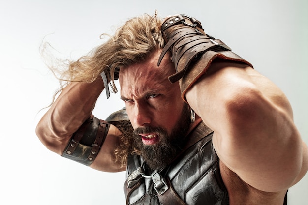 Free photo angry and emotional. blonde long hair and muscular male model in leather viking's costume with the big hammer cosplaying isolated on white studio background. fantasy warrior, antique battle concept.