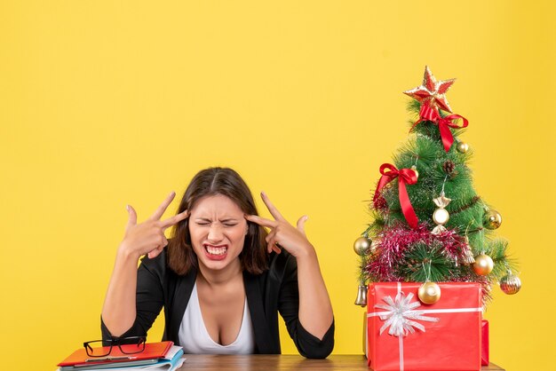 Angry emotional and nervous young woman sitting at a table near decorated Christmas tree at office on yellow 