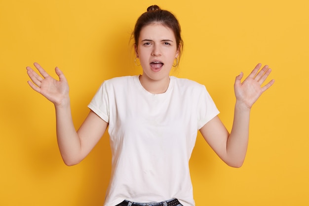 Angry emotional young woman with dark hair and knot, spreading hands aside and screaming