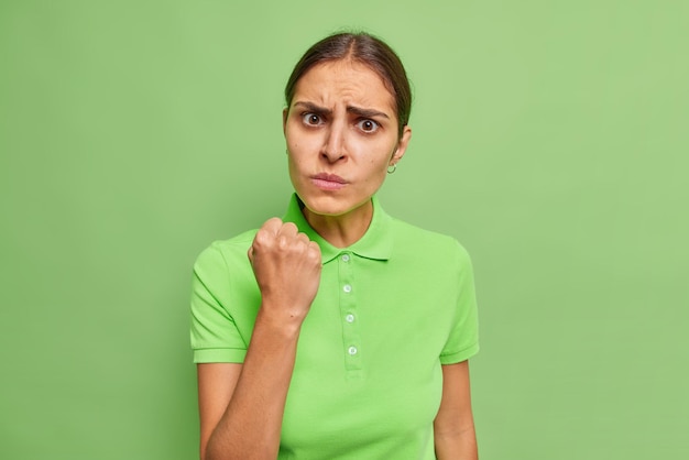 La donna bruna europea arrabbiata ha un'espressione imbronciata che sembra irritata stringe il pugno avverte di qualcosa vestito con una maglietta casual isolata su uno sfondo verde vivido che esprime emozioni negative.