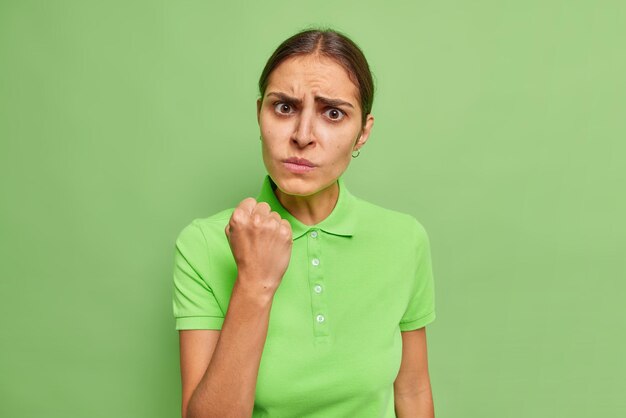화난 유럽 브루네트 여성은 짜증스러운 표정을 짓고 있으며 선명한 녹색 배경 위에 격리된 캐주얼 티셔츠를 입은 무언가에 대해 주먹이 부정적인 감정을 표현한다고 경고합니다.
