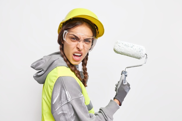 Free photo angry foreman or laborer smirks face holds roller wears protective hard hat transparent glasses involved in home improvement and renovation isolated on white wall. irritated construction worker