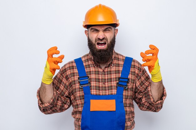 Angry and frustrated bearded builder man in construction uniform and safety helmet wearing rubber gloves shouting and yelling with aggressive expression raising arms going wild