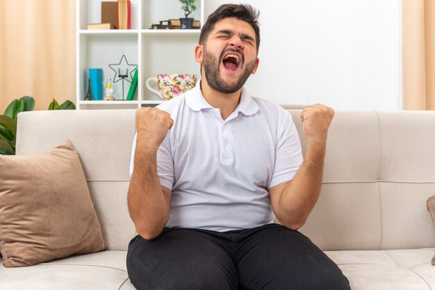 Angry and frustrated young man in casual clothes clenching fists shouting going wild sitting on a couch in light living room