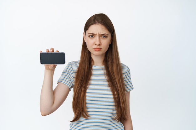 Angry girl frowns, shows mobile phone screen with upset, disappointed face expression, stands in t-shirt on white