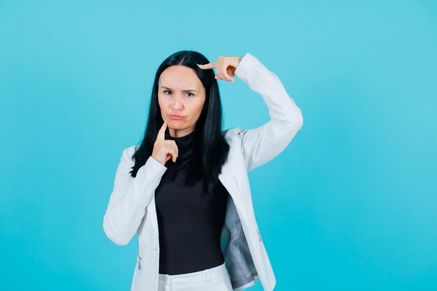 Angry girl is looking at camera by holding forefinger on chin and other on head on blue background