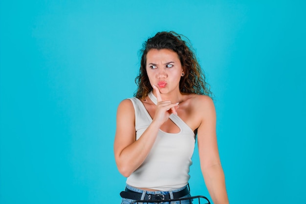 Angry girl is looking left by holding forefiger on chin on blue background
