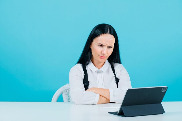 Angry girl is looking at tablet camera by crossing arms on blue background