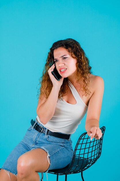 Angry girl is talking on phone by sitting on chair on blue background