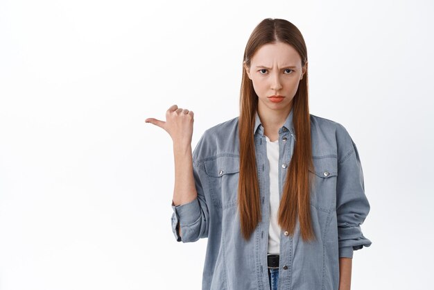 Angry gloomy girl point aside jealous something unfair complaining and looking disappointed sulking and frowning standing displeased against white background