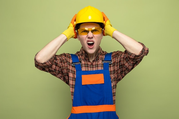 Бесплатное фото Злой схватил голову молодой мужчина-строитель в униформе и перчатках с очками