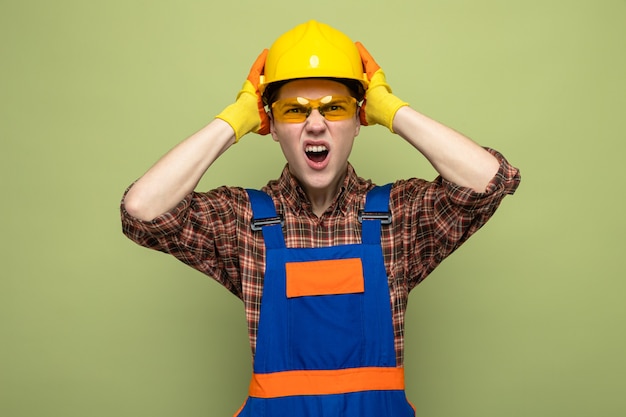 Angry grabbed head young male builder wearing uniform and gloves with glasses 