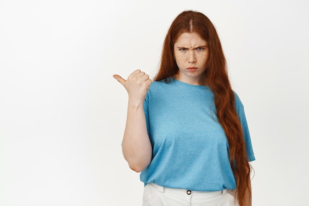 Free photo angry and grumpy redhead girl furrow eyebrows pointing thumb left and frowning displeased complaining sulking about smth unfair standing in tshirt against white background