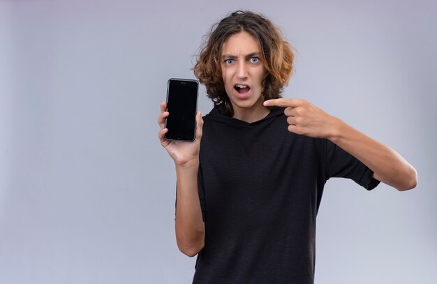 Angry guy with long hair in black t shirt holding a phone and points to phone on white wall