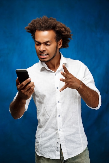 Uomo africano bello arrabbiato che esamina telefono sopra la parete blu.