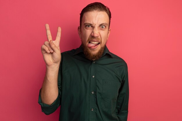 Angry handsome blonde man stucks out tongue and gestures victory hand sign isolated on pink wall