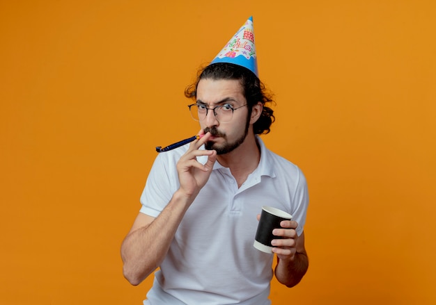 Angry handsome man in birthday cap blowing whistle and holding cup of coffee  isolated on orange background