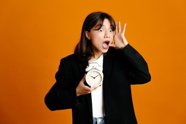 Free photo angry holding alarm clock young beautiful female wearing black jacket isolated on orange background