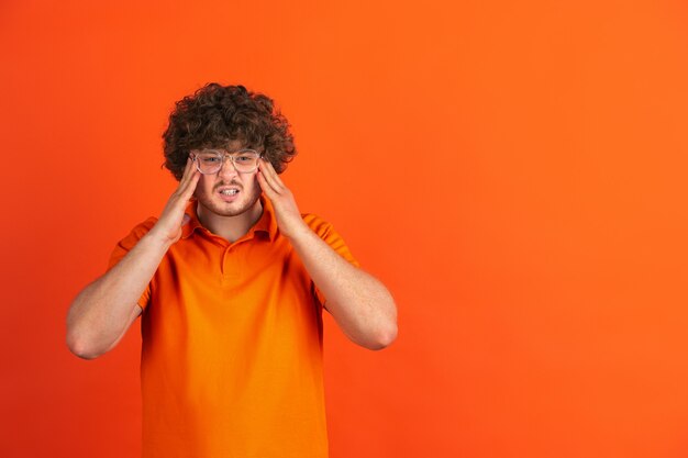 Angry holding head, wtf. Caucasian young man's monochrome portrait on orange  wall. Beautiful male curly model in casual style. 
