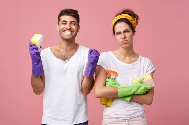 Angry housewife standing crossed hands holding sponge with detergent standing near her happy husband who is rejoicing finishing his work. Couple going to do spring cleaning in their house isolated