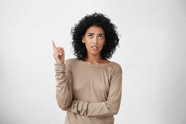 angry and indignant young mixed race female with Afro hairstyle looking up and pointing index finger upwards, feeling mad with noise coming from neighbors above. Body language