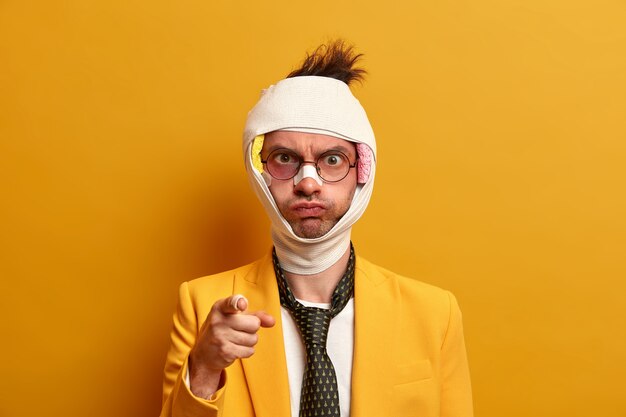 Сердитый раненый госпитализированный мужчина указывает и обвиняет кого-то в своем несчастном случае, у него сотрясение мозга, перевязанная голова, формальная одежда, изолирован на желтой стене, нуждается в лечении