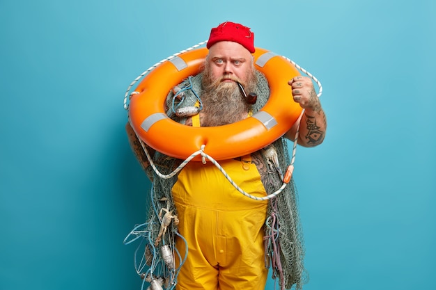Angry irritaed sailor clenches fist, poses with inflated ring, wears red hat and yellow overalls, busy fishing