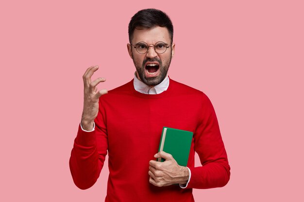 Angry irritated young teacher gestures with hand in annoyance, dressed in red jumper, carries textbook, yells at pupils