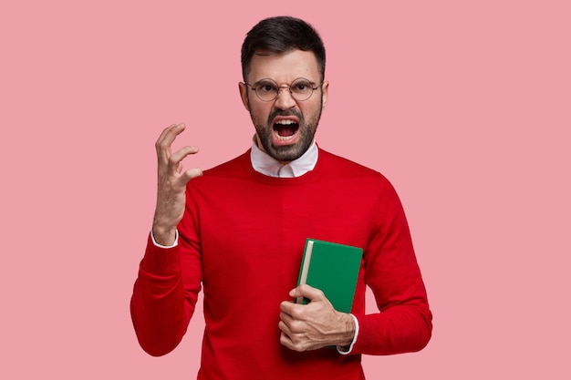 Angry irritated young teacher gestures with hand in annoyance, dressed in red jumper, carries textbook, yells at pupils