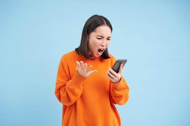 Бесплатное фото Злая корейская девушка, ругающаяся и кричащая на мобильный телефон, выглядит возмущенной и разъяренной, читая новости на