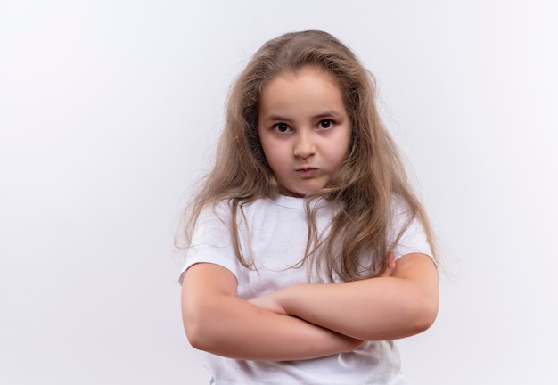 Piccola ragazza arrabbiata della scuola che indossa la maglietta bianca che attraversa le mani sulla parete bianca isolata