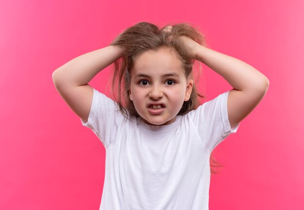 Angry little school girl wearing white t-shirt grabbed head on isolated pink wall