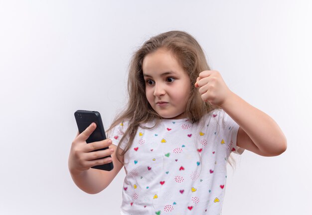 Angry little school woman wearing white t-shirt holding phone and raised fist on isolated white wall