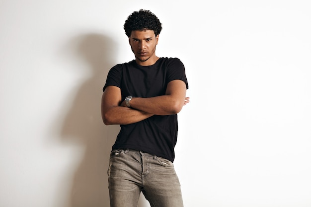 Giovane muscoloso dall'aspetto arrabbiato in t-shirt di cotone nero normale e jeans grigi slim con afro con pareti bianche