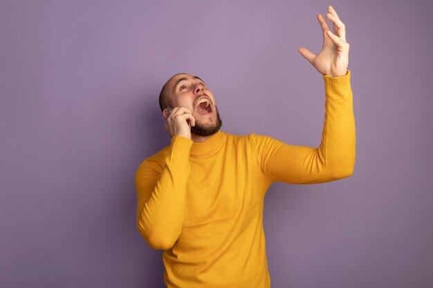 Angry looking up young handsome guy speaks on phone raising hand isolated on purple with copy space