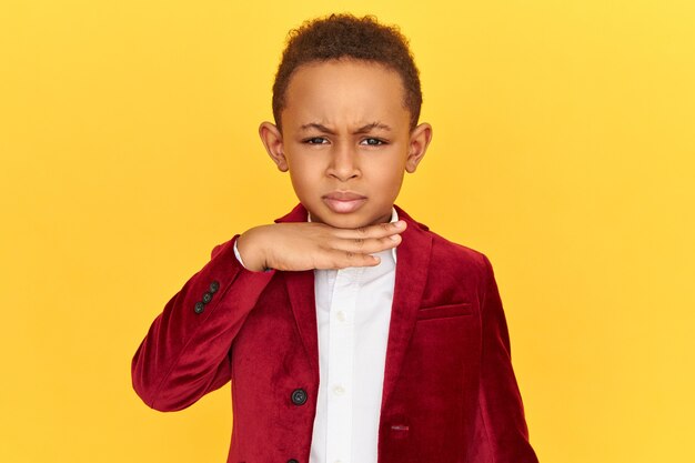 Angry mad dark skinned boy in velvet jacket holding hand on his neck, making menacing gesture, threatening to cut your throat, having mean look.